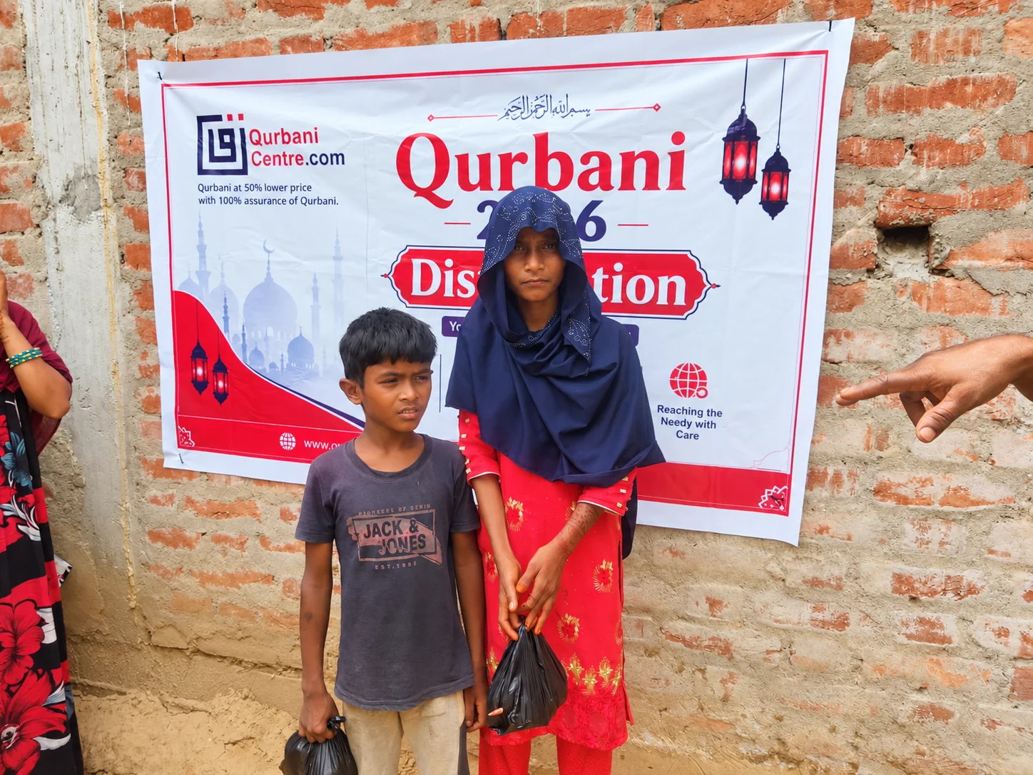 Two children holding small packets stand in front of a Qurbani Centre distribution banner