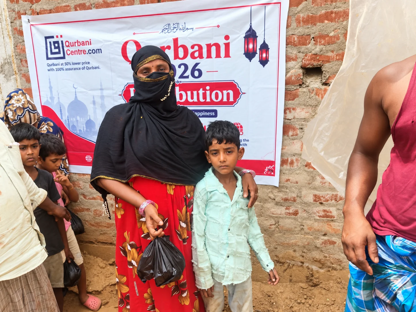 A woman and a boy holding a small packet stand in front of a Qurbani Centre distribution banner